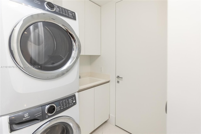 clothes washing area featuring sink, stacked washing maching and dryer, light tile patterned floors, and cabinets