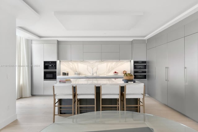 kitchen with a kitchen bar, light hardwood / wood-style flooring, tasteful backsplash, stainless steel double oven, and a raised ceiling