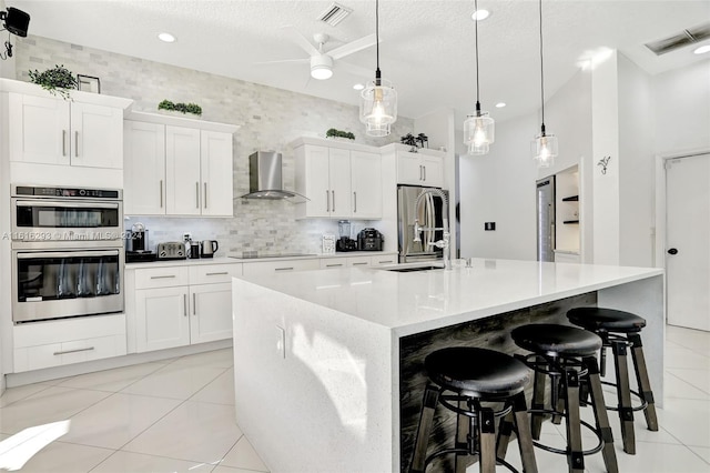 kitchen featuring appliances with stainless steel finishes, an island with sink, light tile patterned floors, white cabinets, and wall chimney exhaust hood