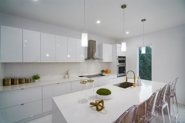 kitchen featuring white cabinetry, wall chimney exhaust hood, an island with sink, and pendant lighting