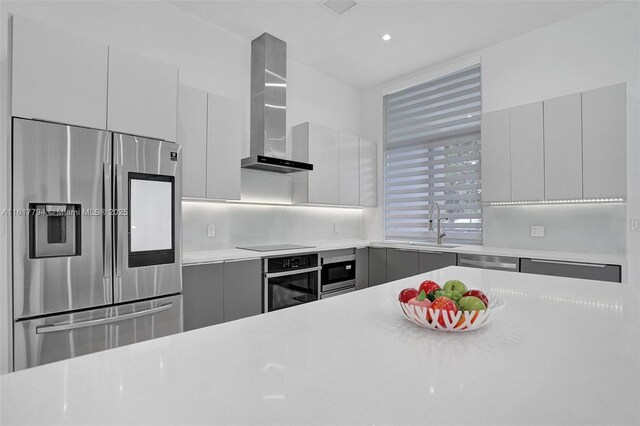 kitchen with gray cabinetry, sink, backsplash, stainless steel appliances, and wall chimney exhaust hood