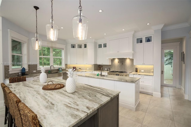 kitchen featuring tasteful backsplash, white cabinets, a kitchen island with sink, light stone countertops, and pendant lighting