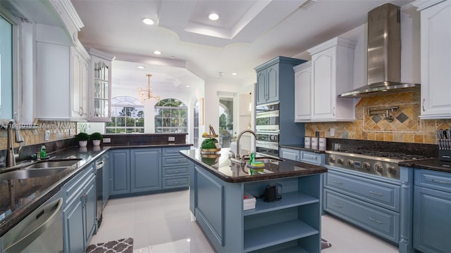 kitchen with wall chimney range hood, an island with sink, sink, blue cabinetry, and appliances with stainless steel finishes