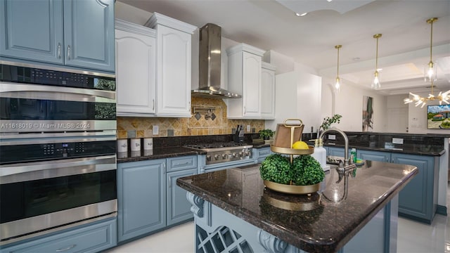 kitchen with appliances with stainless steel finishes, tasteful backsplash, an island with sink, a raised ceiling, and wall chimney exhaust hood