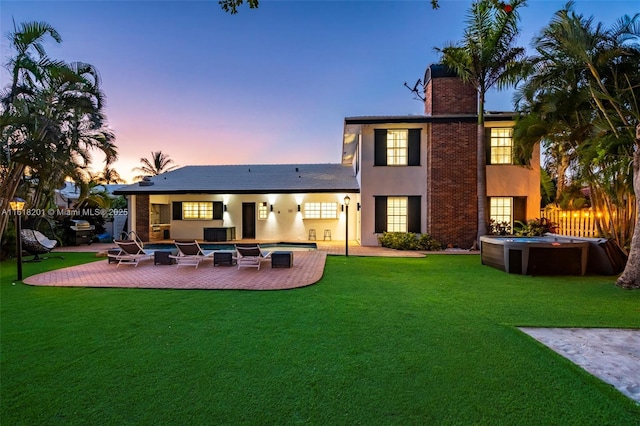 back house at dusk featuring a hot tub, a lawn, and a patio area