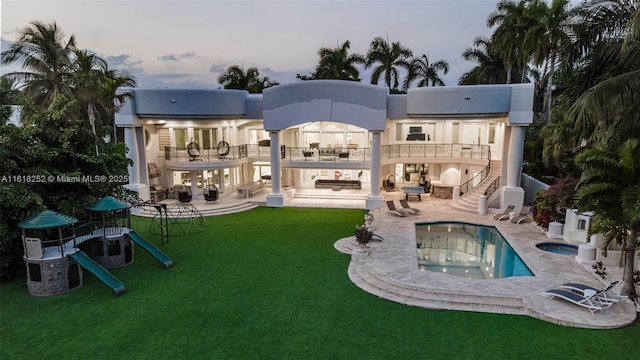 back house at dusk with a playground, a patio, a balcony, a yard, and a pool with hot tub