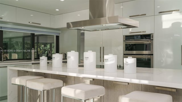 kitchen with white cabinetry, island exhaust hood, a kitchen bar, and double oven