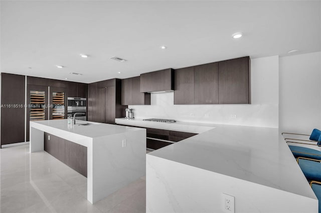 kitchen with double oven, gas stovetop, dark brown cabinets, a center island with sink, and wall chimney exhaust hood