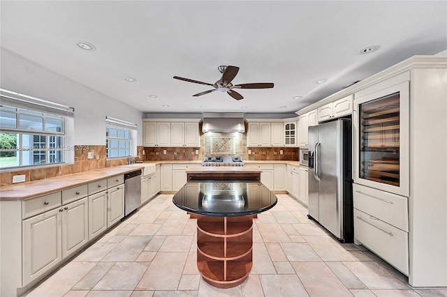 kitchen featuring a center island, wall chimney exhaust hood, stainless steel appliances, decorative backsplash, and light tile patterned floors