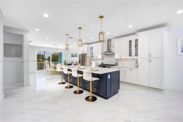 kitchen with white cabinetry, a kitchen breakfast bar, an island with sink, pendant lighting, and wall chimney range hood