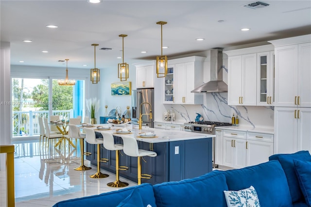 kitchen featuring wall chimney range hood, white cabinetry, stainless steel appliances, a center island with sink, and decorative light fixtures