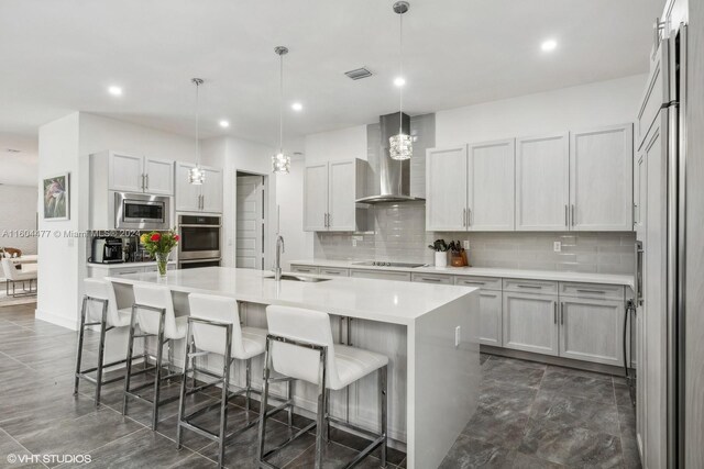 kitchen featuring white cabinetry, wall chimney range hood, built in appliances, pendant lighting, and a kitchen island with sink