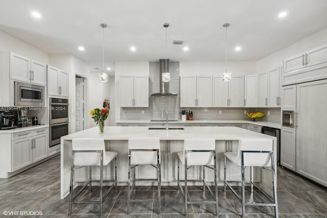 kitchen featuring a kitchen bar, wall chimney exhaust hood, built in appliances, decorative light fixtures, and a center island with sink