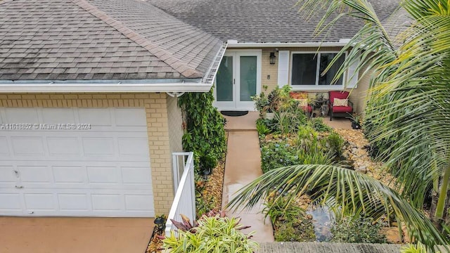 view of exterior entry featuring a shingled roof, brick siding, driveway, and a garage