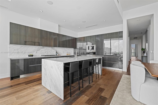 kitchen with wood finished floors, a sink, stainless steel microwave, modern cabinets, and an island with sink