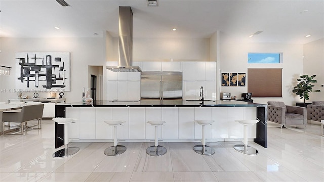 kitchen with sink, wall chimney exhaust hood, built in fridge, a breakfast bar area, and white cabinets