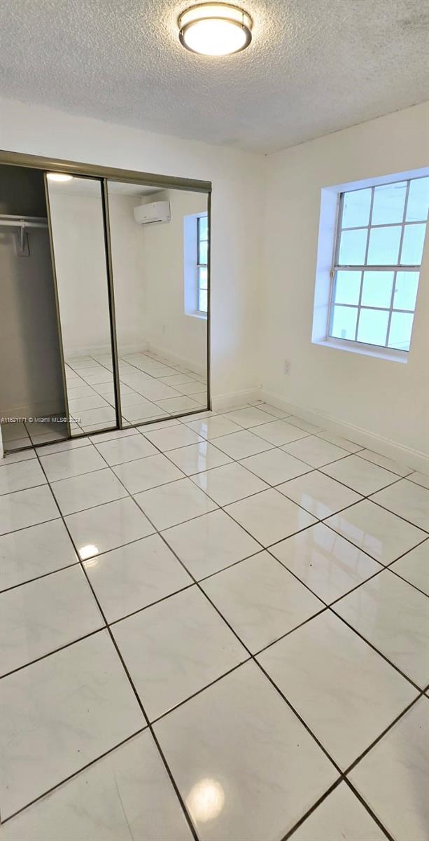 unfurnished bedroom with a wall mounted air conditioner, a textured ceiling, multiple windows, a closet, and light tile patterned floors