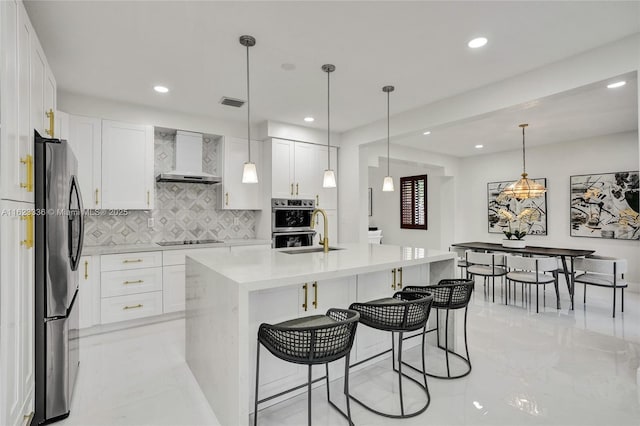 kitchen featuring white cabinetry, hanging light fixtures, a kitchen island with sink, stainless steel appliances, and wall chimney range hood