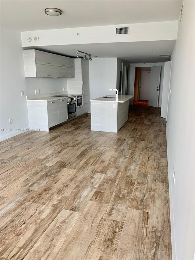 kitchen with sink, white cabinetry, an island with sink, stainless steel oven, and light wood-type flooring