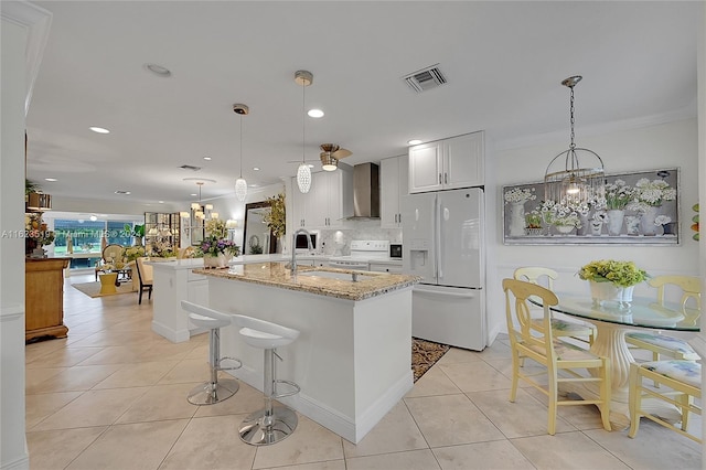 kitchen with white appliances, white cabinets, wall chimney range hood, light stone counters, and backsplash