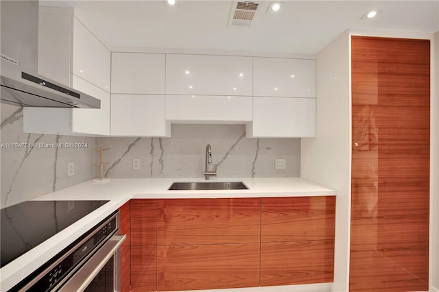 kitchen with tasteful backsplash, white cabinetry, sink, stainless steel oven, and wall chimney range hood