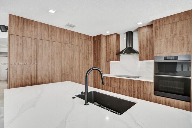 kitchen featuring black electric stovetop, wall chimney exhaust hood, sink, light stone countertops, and double oven