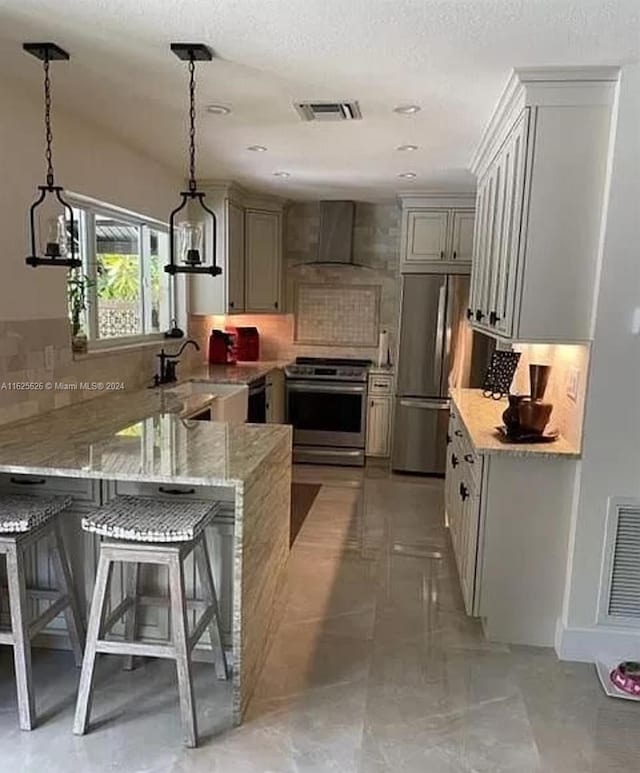 kitchen featuring appliances with stainless steel finishes, backsplash, light stone counters, wall chimney range hood, and decorative light fixtures