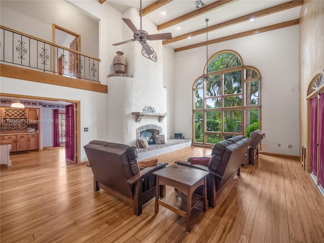 living room with ceiling fan, a high ceiling, beam ceiling, light hardwood / wood-style flooring, and a brick fireplace