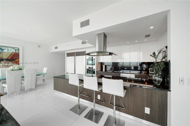 kitchen featuring double oven, white cabinets, island exhaust hood, light tile patterned flooring, and decorative backsplash