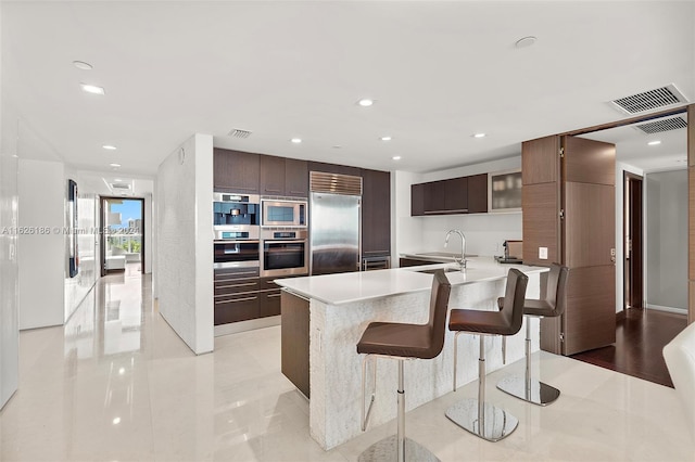 kitchen featuring sink, a breakfast bar, built in appliances, dark brown cabinetry, and an island with sink