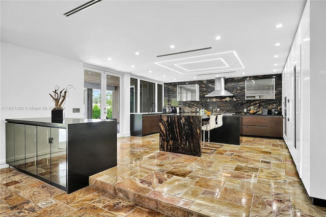 kitchen featuring backsplash, a kitchen bar, sink, wall chimney range hood, and a kitchen island