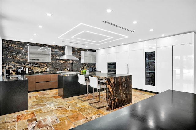 kitchen with white cabinetry, a large island, wall chimney exhaust hood, a kitchen bar, and decorative backsplash