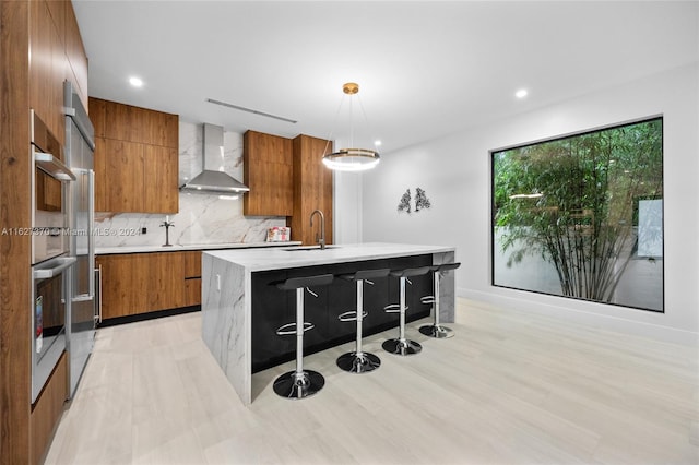 kitchen with a center island with sink, a kitchen breakfast bar, sink, hanging light fixtures, and wall chimney exhaust hood