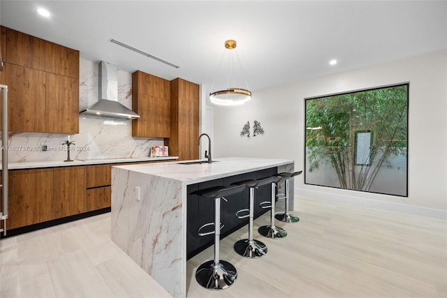 kitchen with a kitchen bar, sink, wall chimney range hood, decorative light fixtures, and a center island with sink