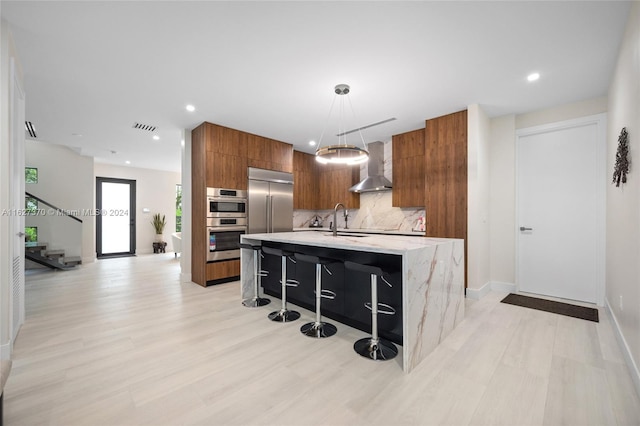 kitchen featuring a kitchen bar, appliances with stainless steel finishes, a kitchen island with sink, wall chimney range hood, and hanging light fixtures