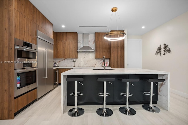 kitchen with sink, wall chimney exhaust hood, an island with sink, decorative light fixtures, and appliances with stainless steel finishes