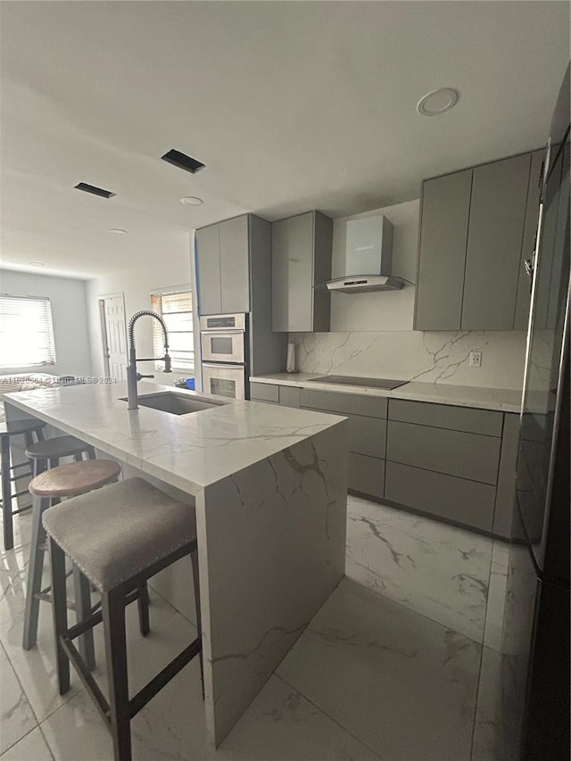 kitchen featuring wall chimney exhaust hood, stainless steel double oven, sink, a center island with sink, and gray cabinets