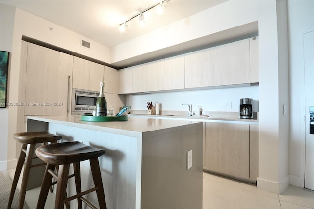 kitchen featuring track lighting, stainless steel microwave, light tile patterned floors, a kitchen breakfast bar, and light brown cabinetry