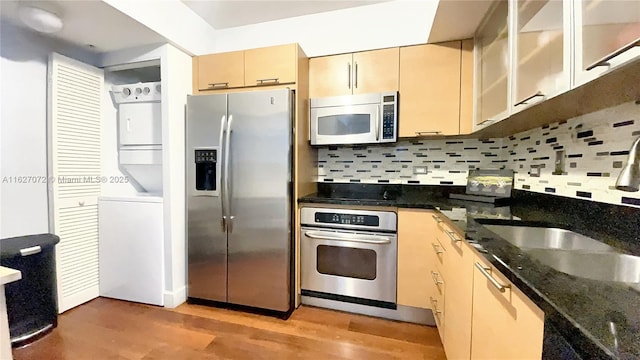 kitchen with stacked washing maching and dryer, stainless steel appliances, backsplash, dark stone countertops, and hardwood / wood-style flooring