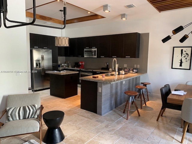 kitchen with a center island, a peninsula, light stone countertops, stainless steel appliances, and dark cabinetry