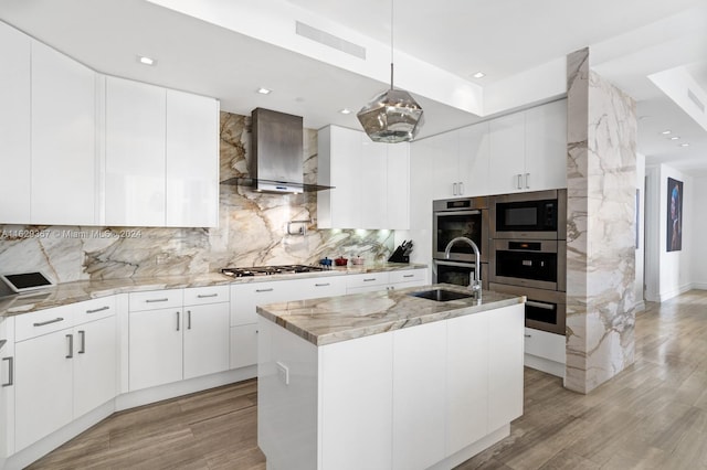 kitchen with wall chimney range hood, backsplash, a kitchen island with sink, appliances with stainless steel finishes, and light hardwood / wood-style floors