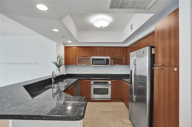 kitchen featuring sink, appliances with stainless steel finishes, a tray ceiling, kitchen peninsula, and dark stone counters