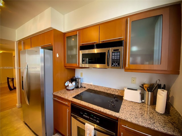 kitchen featuring brown cabinetry, glass insert cabinets, appliances with stainless steel finishes, light stone countertops, and light tile patterned flooring