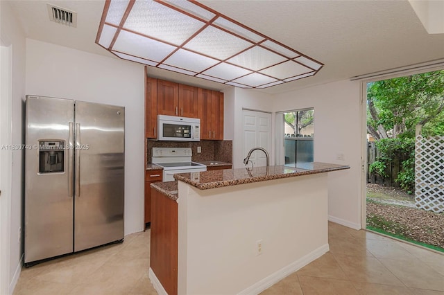 kitchen featuring decorative backsplash, white appliances, light tile patterned floors, a center island with sink, and stone counters