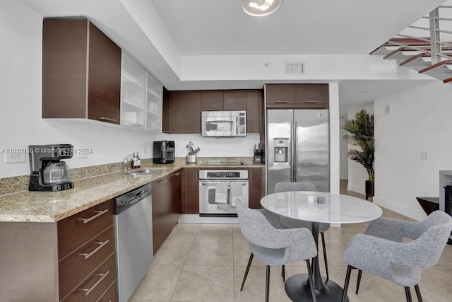 kitchen with dark brown cabinets, appliances with stainless steel finishes, sink, light tile patterned floors, and light stone counters