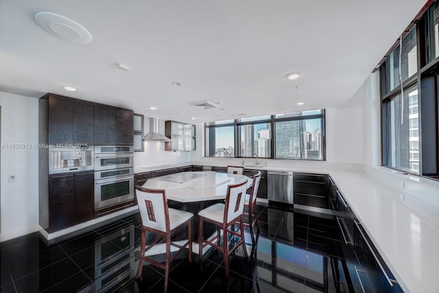 kitchen with appliances with stainless steel finishes, dark brown cabinets, dark tile patterned flooring, kitchen peninsula, and wall chimney exhaust hood