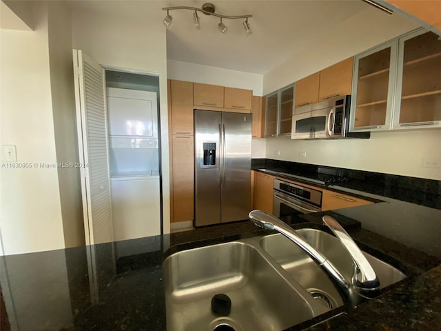 kitchen featuring dark stone countertops, glass insert cabinets, a sink, appliances with stainless steel finishes, and stacked washer / dryer