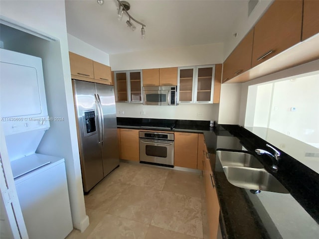 kitchen with glass insert cabinets, stacked washing maching and dryer, dark stone counters, stainless steel appliances, and a sink
