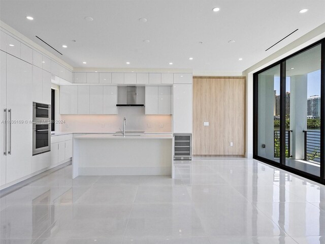 kitchen featuring wall chimney range hood, white cabinets, beverage cooler, and stainless steel double oven