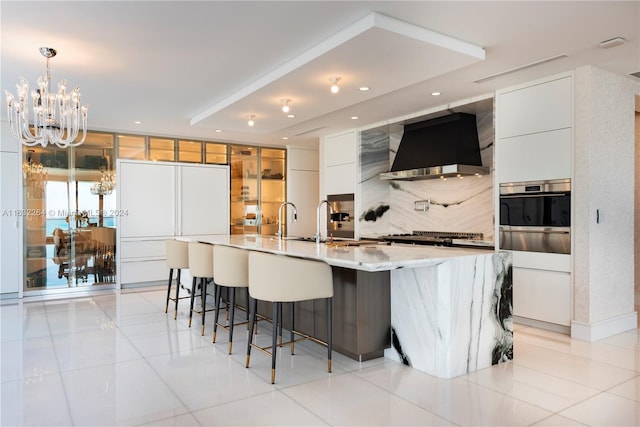 kitchen featuring appliances with stainless steel finishes, white cabinetry, custom range hood, decorative light fixtures, and a large island with sink